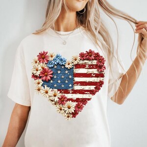 May include: Cream-colored t-shirt featuring a heart-shaped design of the American flag, composed of red, white, and blue flowers. The flag design has a distressed, vintage look, with the stars and stripes clearly visible.