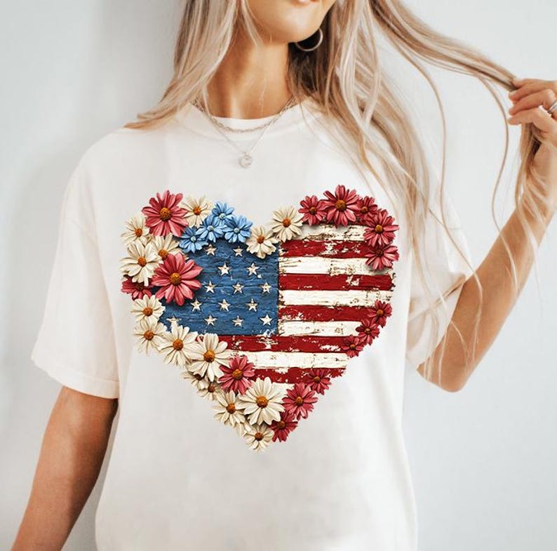 May include: Cream-colored t-shirt featuring a heart-shaped design of the American flag, composed of red, white, and blue flowers. The flag design has a distressed, vintage look, with the stars and stripes clearly visible.