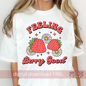 May include: White t-shirt with a pink and red graphic design that says "Feeling Berry Good". The graphic features two strawberries with green leaves and white flowers.