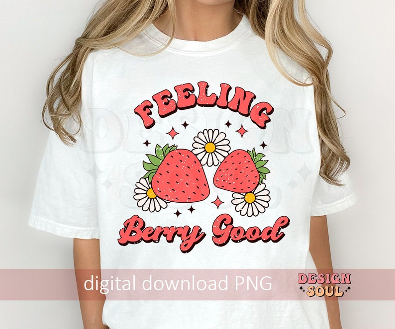 May include: White t-shirt with a pink and red graphic design that says "Feeling Berry Good". The graphic features two strawberries with green leaves and white flowers.
