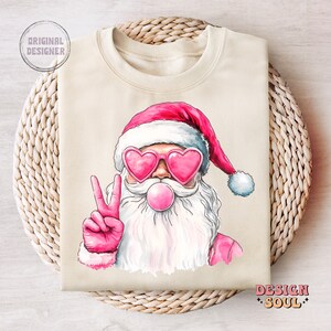 May include: A beige sweatshirt featuring a pink and white illustration of Santa Claus wearing heart-shaped sunglasses and blowing a bubble. Santa is giving the peace sign with his right hand.