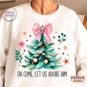 May include: White sweatshirt with a green Christmas tree with pink bow and gold ornaments. The text "Oh come, let us adore Him" is printed below the tree.