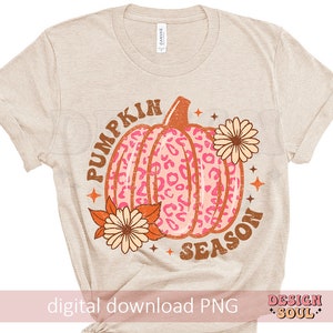 May include: A beige t-shirt with the text "Pumpkin Season" in brown and orange letters. A pink and orange pumpkin with a leopard print design is in the center of the text. There are two orange and yellow flowers on either side of the pumpkin.