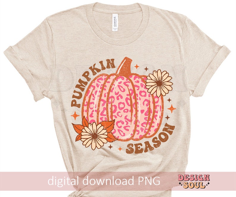 May include: A beige t-shirt with the text "Pumpkin Season" in brown and orange letters. A pink and orange pumpkin with a leopard print design is in the center of the text. There are two orange and yellow flowers on either side of the pumpkin.