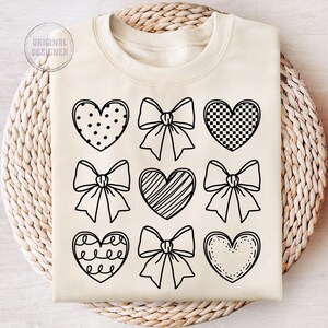 May include: A cream-colored sweatshirt with a black outline of various hearts and bows. The hearts have different patterns, including polka dots, checkered, and stripes.