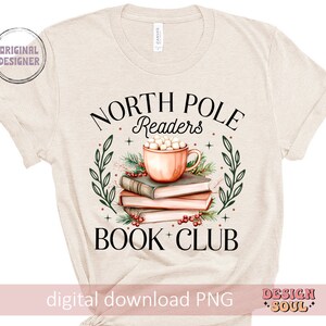 May include: A beige t-shirt with the text "North Pole Readers Book Club" in black. The design features a mug of hot chocolate with marshmallows, surrounded by books and greenery.