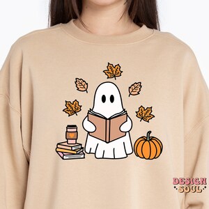 May include: A tan sweatshirt with a white ghost reading a book, surrounded by orange leaves and a pumpkin. The ghost is holding a cup of coffee. The text "DESIGN SOUL" is printed on the bottom right corner of the sweatshirt.