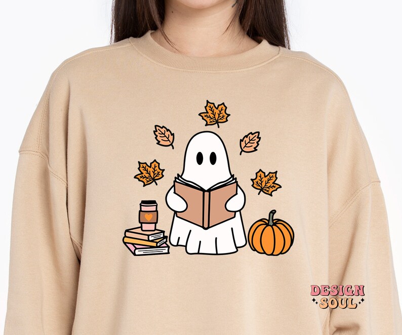 May include: A tan sweatshirt with a white ghost reading a book, surrounded by orange leaves and a pumpkin. The ghost is holding a cup of coffee. The text "DESIGN SOUL" is printed on the bottom right corner of the sweatshirt.