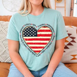 May include: A light blue t-shirt with a distressed American flag heart design. The heart is surrounded by a circle of text that reads: *Victory* *USA* *Freedom* *Independence* *Liberty* *Patriotism* *Unity* *Home* *Land of the free* *America*.