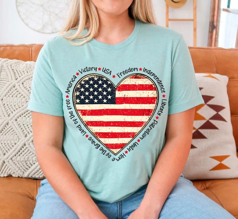 May include: A light blue t-shirt with a distressed American flag heart design. The heart is surrounded by a circle of text that reads: *Victory* *USA* *Freedom* *Independence* *Liberty* *Patriotism* *Unity* *Home* *Land of the free* *America*.