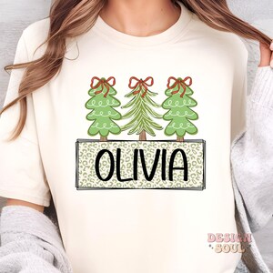 May include: A white t-shirt with a design of three green Christmas trees with red bows and the name "Olivia" in a leopard print box.
