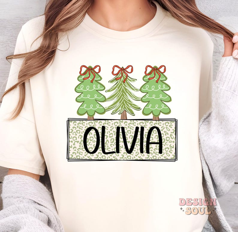 May include: A white t-shirt with a design of three green Christmas trees with red bows and the name "Olivia" in a leopard print box.