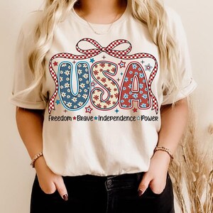 May include: A white t-shirt with a red, white, and blue USA graphic. The graphic features a bow with stars and flowers. The text "Freedom Brave Independence Power" is printed below the graphic.
