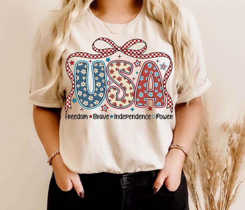 May include: A white t-shirt with a red, white, and blue USA graphic. The graphic features a bow with stars and flowers. The text "Freedom Brave Independence Power" is printed below the graphic.