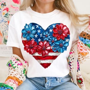 May include: White t-shirt with a red, white, and blue heart-shaped design. The heart is made up of flowers with the American flag colors.