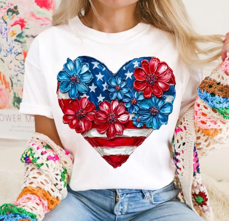 May include: White t-shirt with a red, white, and blue heart-shaped design. The heart is made up of flowers with the American flag colors.