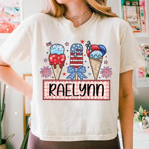 May include: A white t-shirt with a red, white, and blue ice cream cone design. The ice cream cones are decorated with stars and stripes. The text "Raelynn" is written in a red and white checkered box.