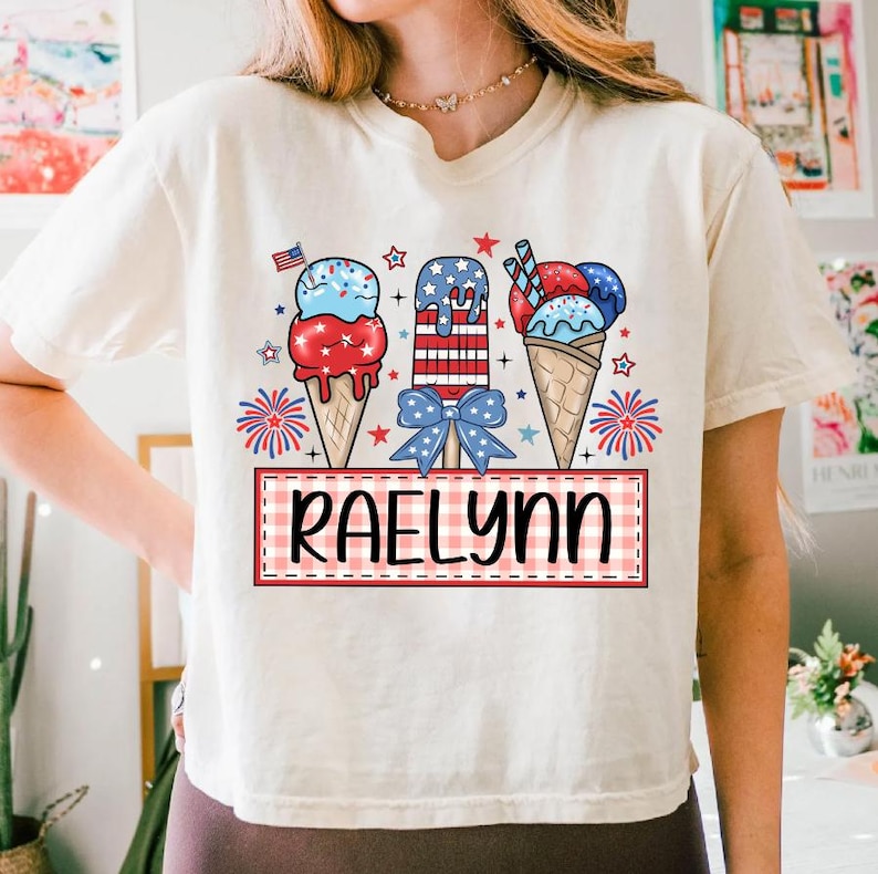 May include: A white t-shirt with a red, white, and blue ice cream cone design. The ice cream cones are decorated with stars and stripes. The text "Raelynn" is written in a red and white checkered box.