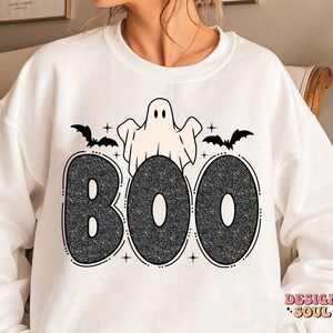 May include: White sweatshirt with a black glitter graphic of the word "BOO" with a ghost and bats. The ghost is white with black eyes and a black outline. The bats are black with white outlines. The word "BOO" is in a black glitter font.