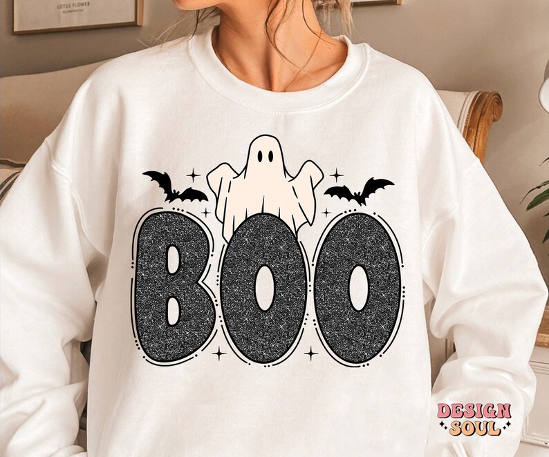 May include: White sweatshirt with a black glitter graphic of the word "BOO" with a ghost and bats. The ghost is white with black eyes and a black outline. The bats are black with white outlines. The word "BOO" is in a black glitter font.