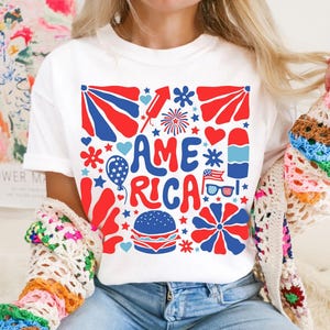 以下が含まれることがあります： 星、ハート、花火、その他の愛国的なシンボルで囲まれた「AMERICA」という言葉が描かれた、赤、白、青のグラフィックデザインが施された白いTシャツです。