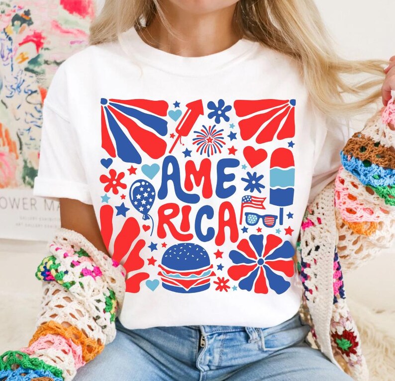 以下が含まれることがあります： 星、ハート、花火、その他の愛国的なシンボルで囲まれた「AMERICA」という言葉が描かれた、赤、白、青のグラフィックデザインが施された白いTシャツです。