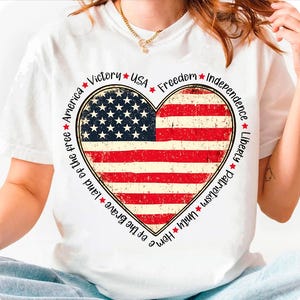 May include: White t-shirt with a red, white, and blue heart-shaped American flag design. The text around the heart reads "Home of the Brave Land of the free America Victory USA Freedom Independence Liberty Patriotism Unity".