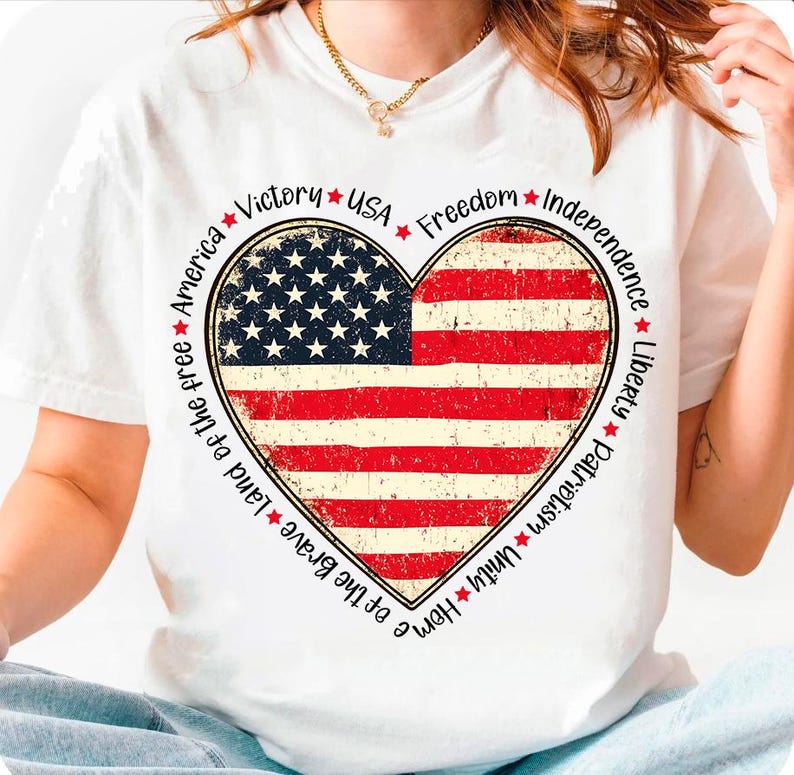 May include: White t-shirt with a red, white, and blue heart-shaped American flag design. The text around the heart reads "Home of the Brave Land of the free America Victory USA Freedom Independence Liberty Patriotism Unity".