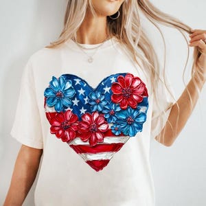 May include: A white t-shirt with a red, white, and blue heart-shaped design. The heart is made up of flowers and the American flag.