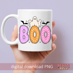 Op de afbeelding: Een witte keramische mok met een spook- en vleermuisontwerp. De mok heeft het woord "BOO" erop geschreven in roze, oranje en roze stippen.