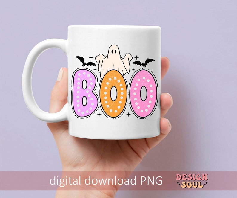 Op de afbeelding: Een witte keramische mok met een spook- en vleermuisontwerp. De mok heeft het woord "BOO" erop geschreven in roze, oranje en roze stippen.