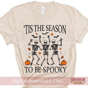 May include: A beige t-shirt with the text "'Tis the season to be spooky" printed on it. The text is in black and there are three skeletons dancing with pumpkins in front of them.