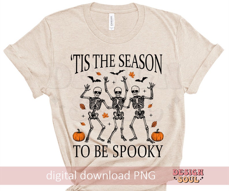 May include: A beige t-shirt with the text "'Tis the season to be spooky" printed on it. The text is in black and there are three skeletons dancing with pumpkins in front of them.