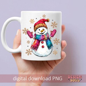 May include: White ceramic mug with a colorful snowman illustration. The snowman is wearing a red hat, a rainbow scarf, and mittens. The background is decorated with gold snowflakes.