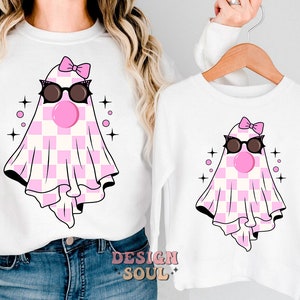 May include: A white long-sleeved shirt with a pink and white checkered ghost wearing sunglasses and a pink bow. The ghost has a pink bubble gum in its mouth. The shirt has the text "DESIGN SOUL" on the bottom.