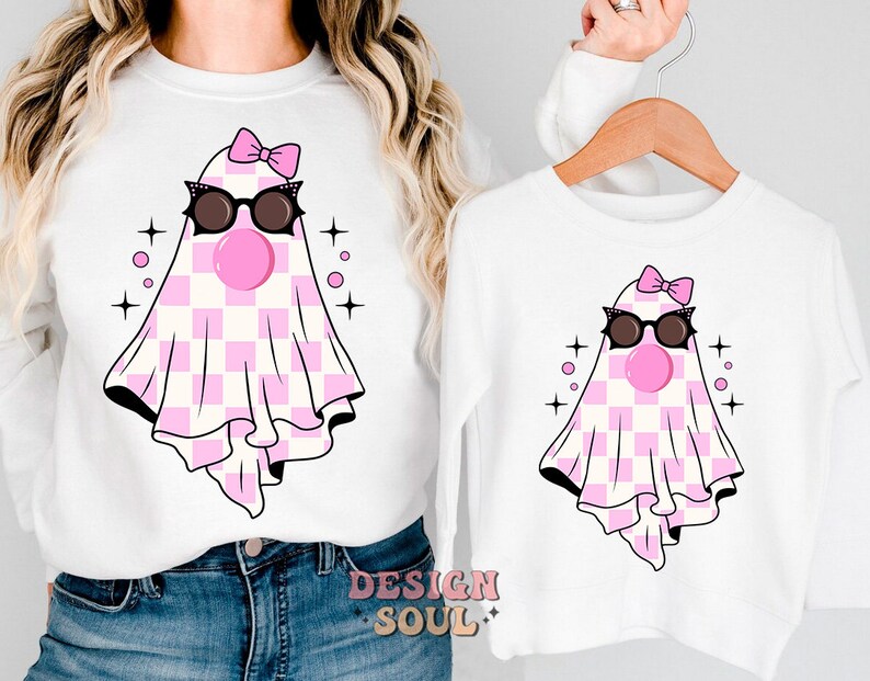 May include: A white long-sleeved shirt with a pink and white checkered ghost wearing sunglasses and a pink bow. The ghost has a pink bubble gum in its mouth. The shirt has the text "DESIGN SOUL" on the bottom.