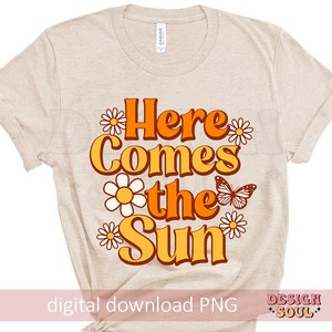May include: A beige t-shirt with a graphic design that says "Here Comes the Sun" in orange and yellow letters with a butterfly and flowers in orange, yellow, and white.