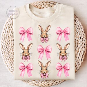May include: A beige sweatshirt with a repeating pattern of pink bows and brown bunnies wearing pink bows. The bunnies have brown eyes and noses.