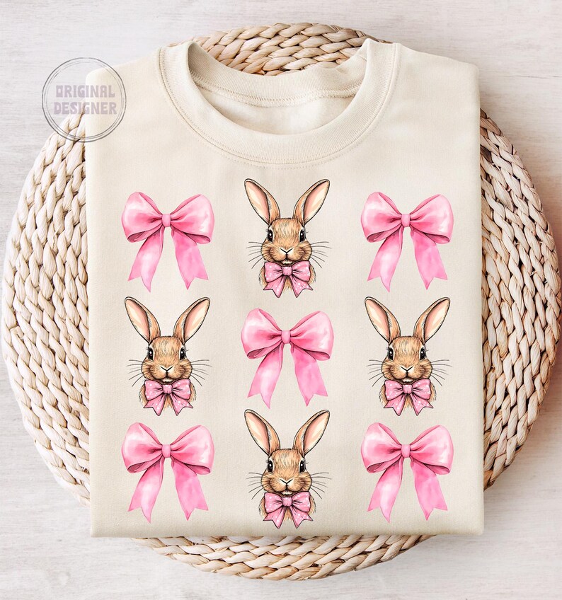 May include: A beige sweatshirt with a repeating pattern of pink bows and brown bunnies wearing pink bows. The bunnies have brown eyes and noses.