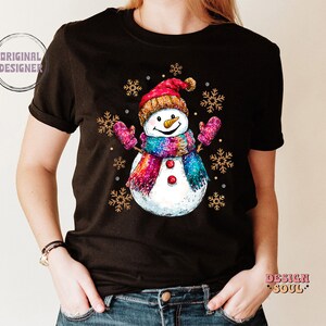 May include: Black t-shirt with a colorful snowman wearing a red hat and a pink scarf. The snowman is surrounded by snowflakes.
