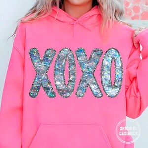 May include: A pink hoodie with a large iridescent glitter XOXO graphic on the front. The hoodie has a drawstring hood and a kangaroo pocket.
