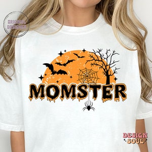 May include: White t-shirt with a black and orange Halloween-themed graphic. The graphic features a spooky orange moon with bats, a spider web, and a tree. The text "MOMSTER" is written in black dripping letters.
