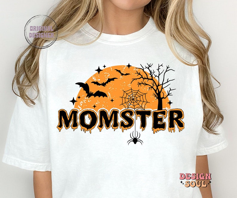 May include: White t-shirt with a black and orange Halloween-themed graphic. The graphic features a spooky orange moon with bats, a spider web, and a tree. The text "MOMSTER" is written in black dripping letters.