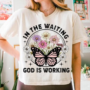 May include: White t-shirt with a floral design and a butterfly. The text "In the waiting God is working" is printed on the shirt.