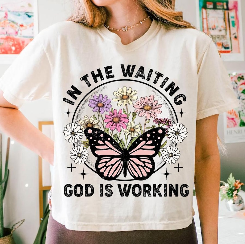 May include: White t-shirt with a floral design and a butterfly. The text "In the waiting God is working" is printed on the shirt.