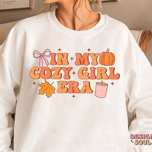 Puede incluir: Sudadera blanca con el texto "In My Cozy Girl Era" en letras naranja y rosa. El dise&ntilde;o tambi&eacute;n incluye una calabaza, una vela, un lazo y una hoja.