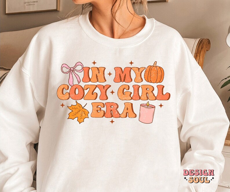 Puede incluir: Sudadera blanca con el texto "In My Cozy Girl Era" en letras naranja y rosa. El dise&ntilde;o tambi&eacute;n incluye una calabaza, una vela, un lazo y una hoja.