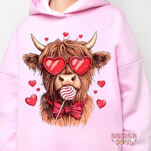 May include: Pink hoodie with a graphic of a Highland cow wearing heart-shaped sunglasses and holding a lollipop. The cow is wearing a red bow tie. The text "DESIGN SOUL" is printed on the bottom right corner of the hoodie.