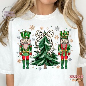 May include: White t-shirt with a Christmas design. It features two nutcrackers, a green Christmas tree, and a black and white polka dot bow. The design includes snowflakes and the words "Design Soul" and "Original Designer".