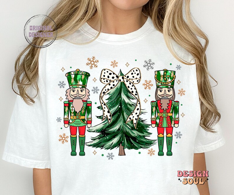 May include: White t-shirt with a Christmas design. It features two nutcrackers, a green Christmas tree, and a black and white polka dot bow. The design includes snowflakes and the words "Design Soul" and "Original Designer".